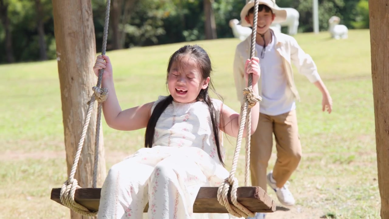 Una bambina si dondola su un'altalena di legno mentre suo fratello la guarda in un parco.