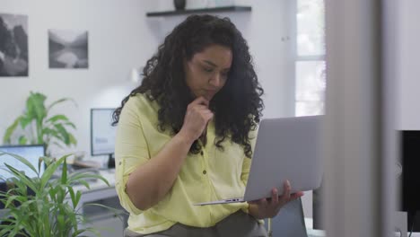 Thoughtful-biracial-businesswoman-working,-using-laptop-holding-her-chin-in-modern-office