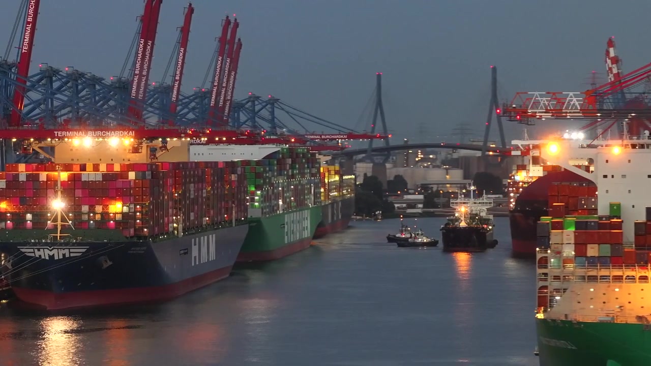 Telefoto-udsigt over Hamborg Havn med store containerskibe og tårnhøje lastkraner, der skaber lagdelt perspektiv og bevægelse i skumringen.