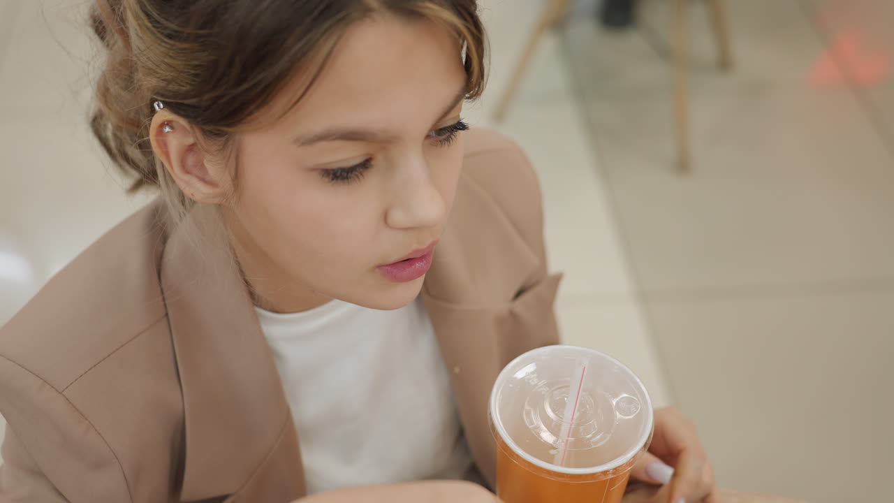 쇼핑몰 테이블에서 음료를 마시는 십 대 백인 학생, 빨대가 꽂힌 오렌지색 컵, 블레이저 의상, 생각에 잠긴 표정, 컵에 올려진 편안한 손, 실내 푸드 코트 배경, 부드러운 햇빛, 조용한 휴식