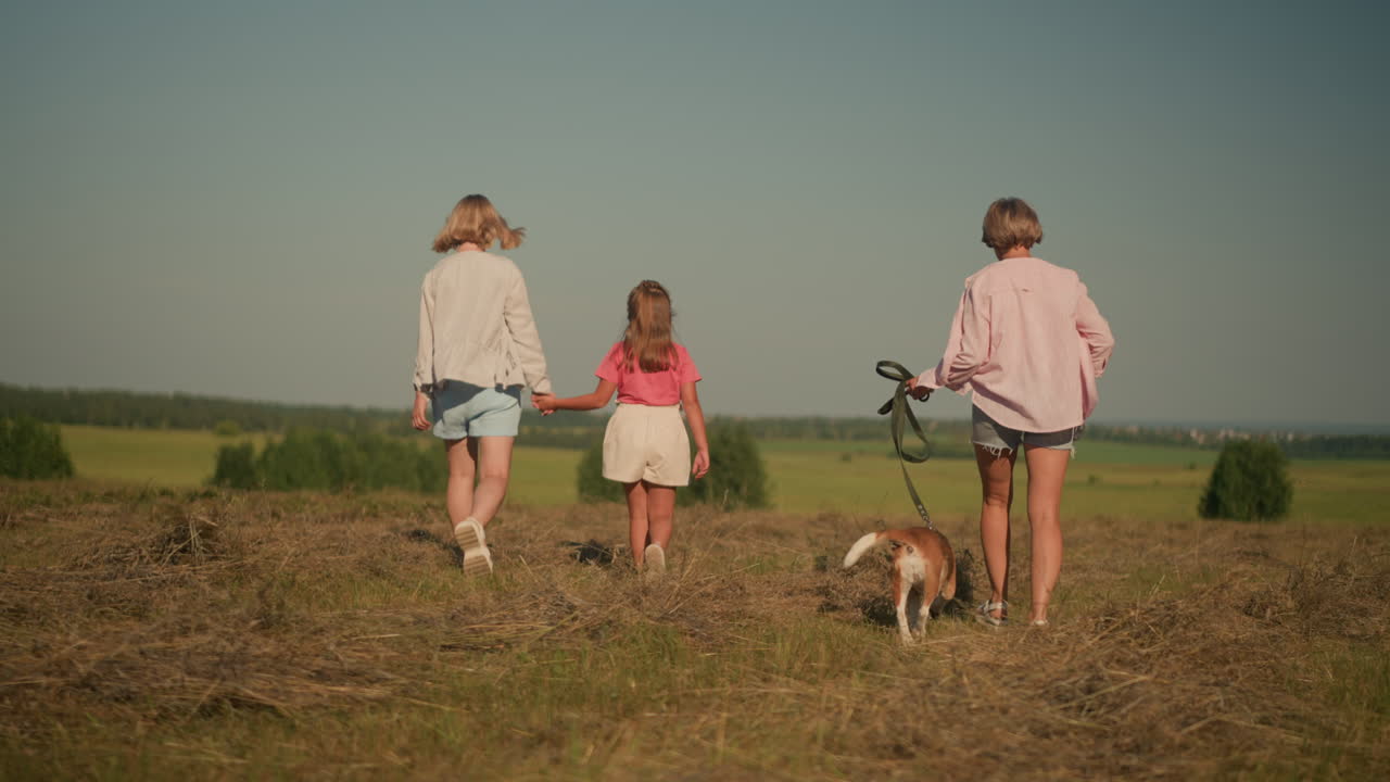 обратный вид любительницы домашних животных, гуляющей с собакой на поводке и матерью, держащей дочь за руку, когда они прогуливаются по сельскохозяйственной земле, ее волосы дуют на ветру, окружены пышной зеленью и далекими деревьями