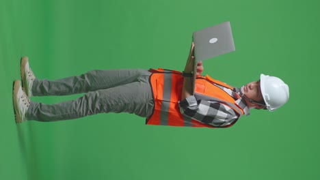 full body side view of asian male engineer with safety helmet working on a laptop while standing in the green screen background studio