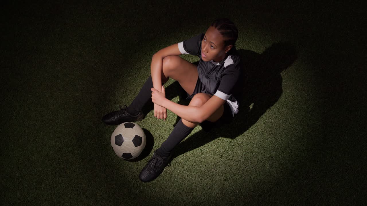 Riflettore sul pallone che attira una donna africana adulta in tenuta sportiva, la quale si avvicina e si siede, guardando la luce sull'erba sintetica.