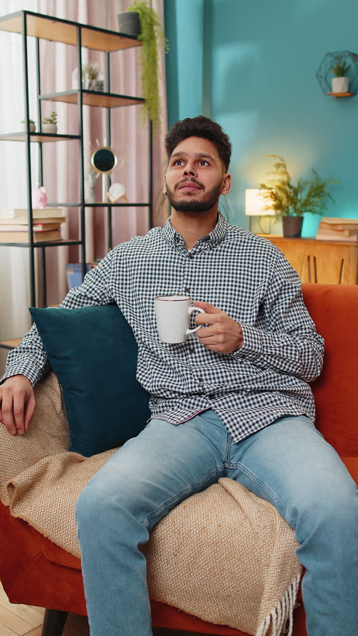 atractivo joven indio sonriente bebiendo una taza de café o té de hierbas en el sofá de casa por la mañana