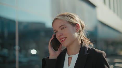 young lady in professional attire with warm smile on phone call walking by glass building under bright sunlight, city reflections visible on glass, radiating positivity and calmness in urban environment