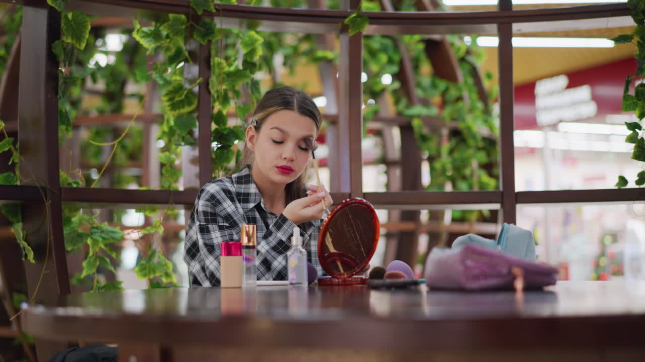 젊은 소녀는 메이크업 브러쉬를 사용하여 눈 위에 파우더를 적용하고, 메이크업 도구와 거울을 보면서 테이블에 어져있는 재료와 블러쉬와 같은 화장품 제품