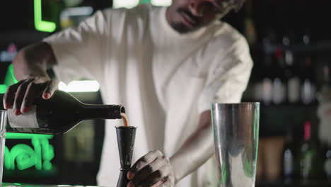 bartender pouring liquor from bottle into jigger for cocktail preparation at bar counter, with focus on precision and vibrant green neon lighting in background