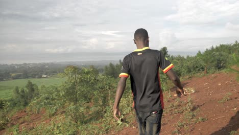 Una-Toma-En-Cámara-Lenta-Desde-Atrás-De-Un-Joven-Africano-Mientras-Camina-Bajo-El-Brillo-Del-Sol-De-La-Mañana-En-Un-Camino-De-Arbustos-En-África