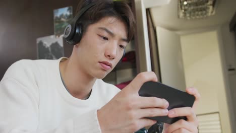 Asian-boy-wearing-headphones-playing-game-on-smartphone-sitting-on-the-couch-at-home