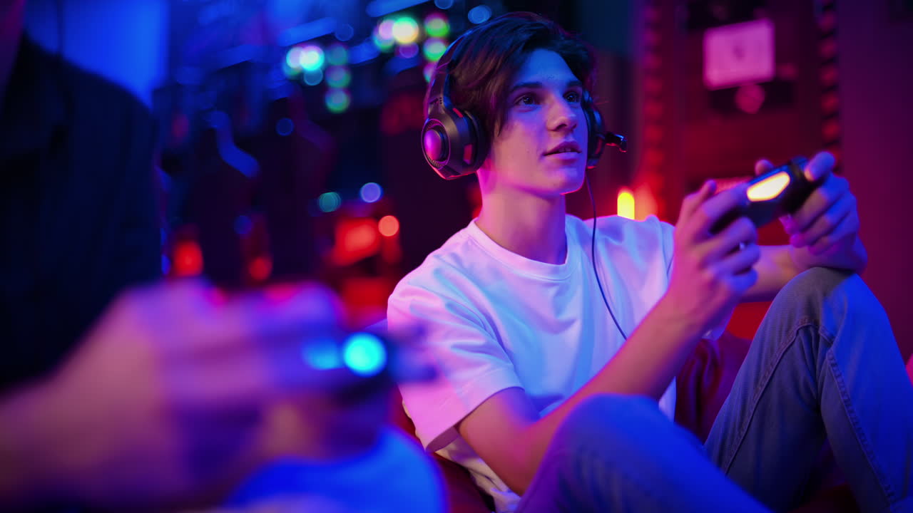 Two teen friends in headphones are talking and playing a game console using gamepads, talking and laughing while sitting on bean bags. Blue and red illumination