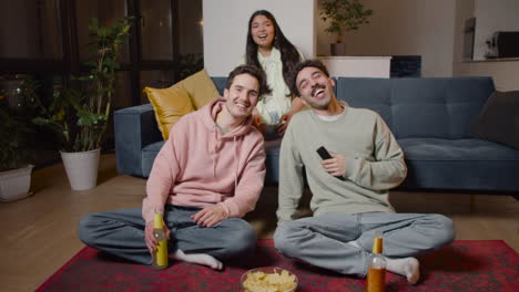 three friends watching comic movie on television sitting on couch and floor, eating popcorn and chips and drinking soda