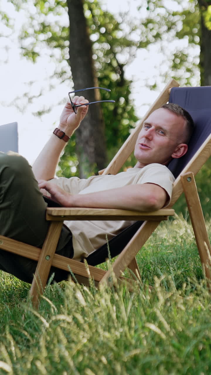 Man took a break from work and laid back in folding chair. Freelancer holds laptop and looks aside speaking, searching for ideas. Vertical video