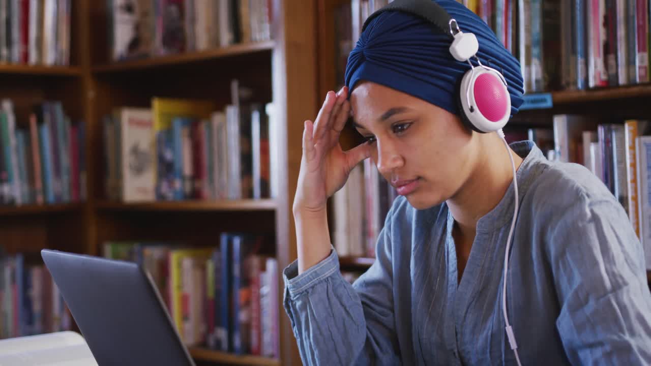 estudiante asiática vestida con un hijab azul sentada y escuchando música en la biblioteca