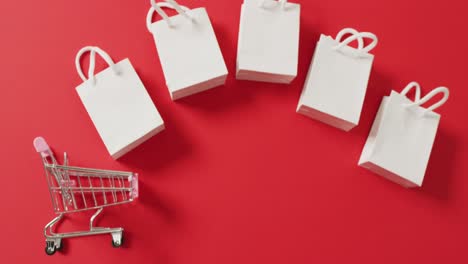 cinco bolsas de regalos blancas y un carrito de compras sobre un fondo rojo con espacio de copia