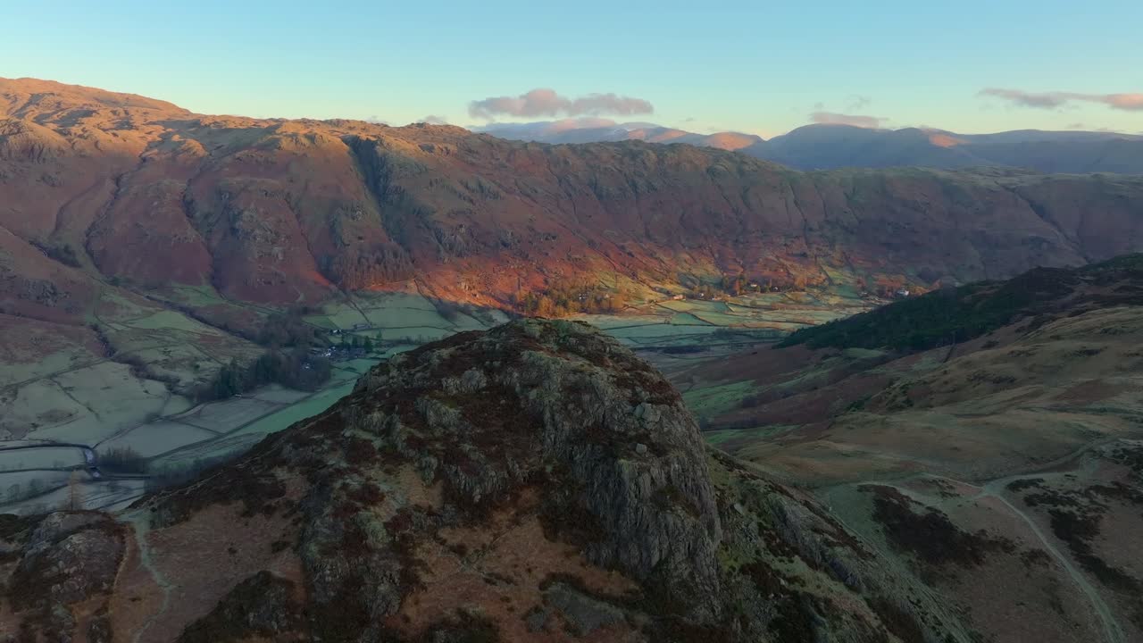 Flying over craggy mountaintop revealing wintry valley with mountain peaks bathed in winter dawn light. Langdales, English Lake District, Cumbria, UK.