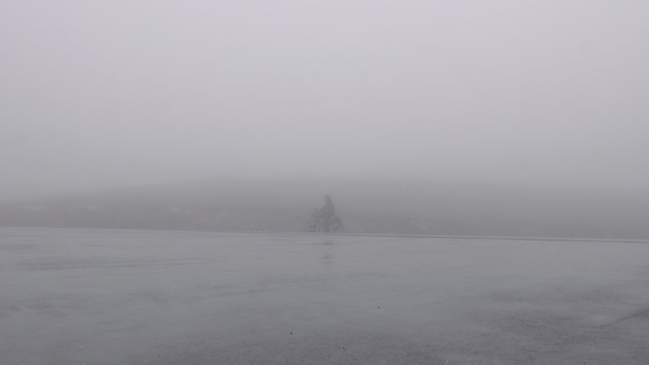 En vuxen cyklist cyklar genom tät dimma när vägen försvinner i nästan obefintlig sikt längs Kvænangsveien, Kvænangsfjellet, Norge – statisk bild.