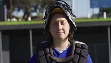 portrait of caucasian female baseball player, catcher, in protective clothing, smiling on field