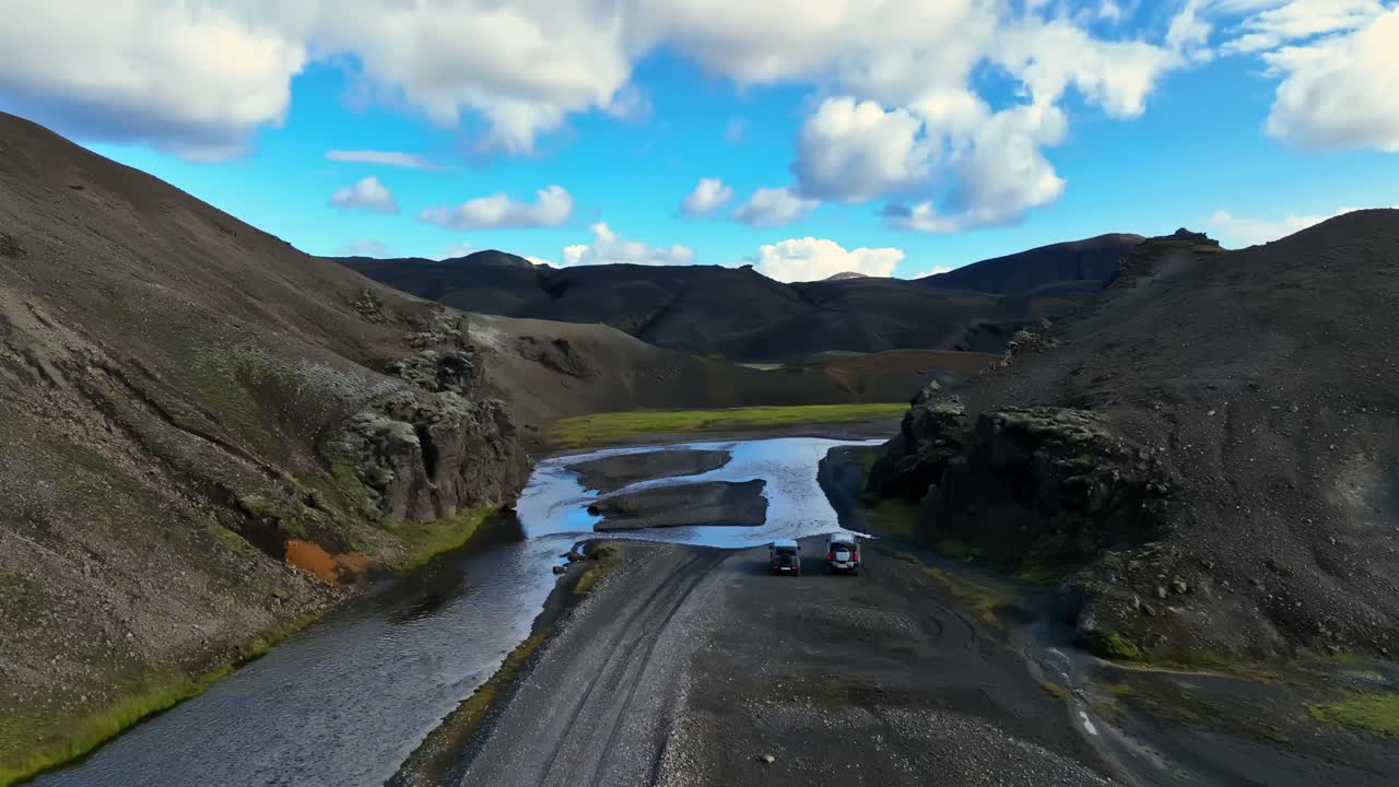 Two 4×4 vehicles navigate a volcanic river crossing in Iceland’s highlands, one moving and one parked on the gravel bank, surrounded by dark cliffs, clear sky, and branching water channels