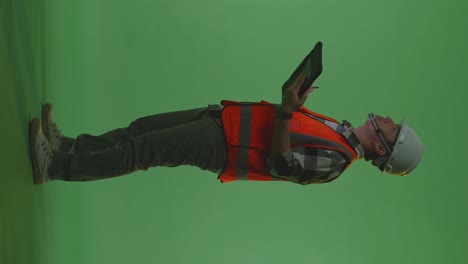 full body side view of asian male engineer with safety helmet taking note on the tablet and looking around while standing in the green screen background studio, during sunset or sunrise time