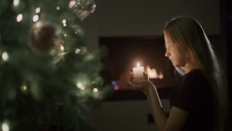 A-girl-with-a-candle-in-her-hands-sits-by-the-Christmas-tree.-In-the-background,-a-fire-burns-in-a-fireplace.-Christmas-Eve