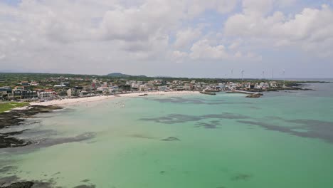 Stunning-drone-view-of-Woljeong-Beach's-pristine-shores,-a-tranquil-oasis-on-beautiful-Jeju-Island,-with-a-unique-view-of-Wind-Turbines-in-right-on-the-waters-edge