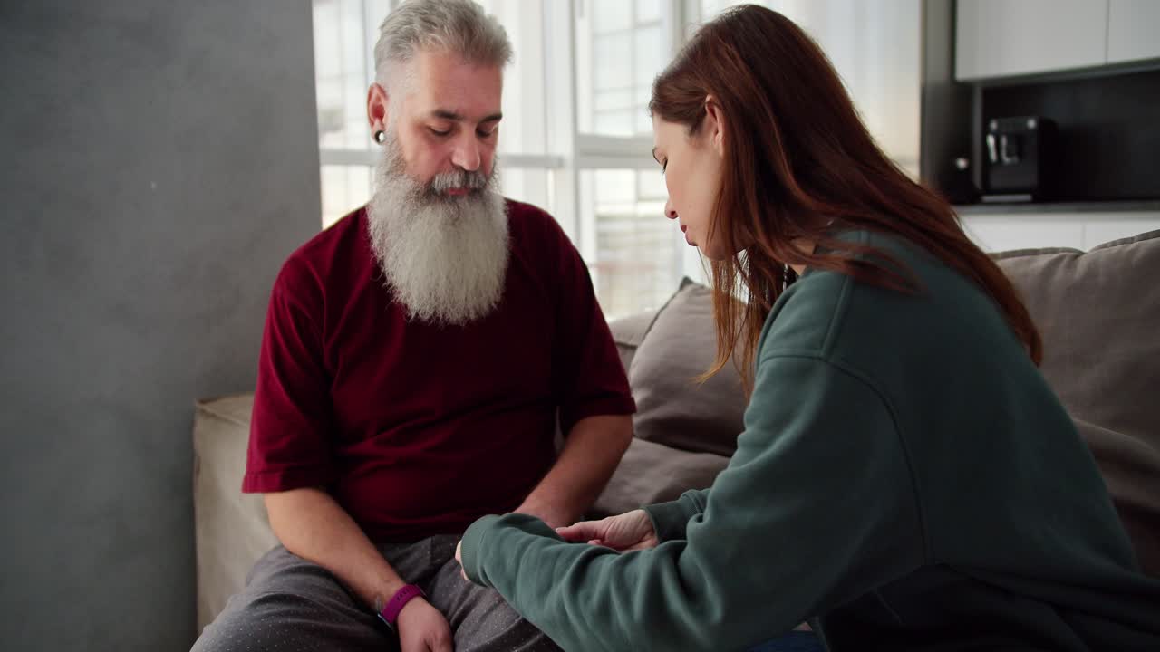 A brunette girl in a green sweater measures the pulse of her elderly father with gray hair and a lush beard in a red T-shirt while sitting on a modern sofa in the apartment