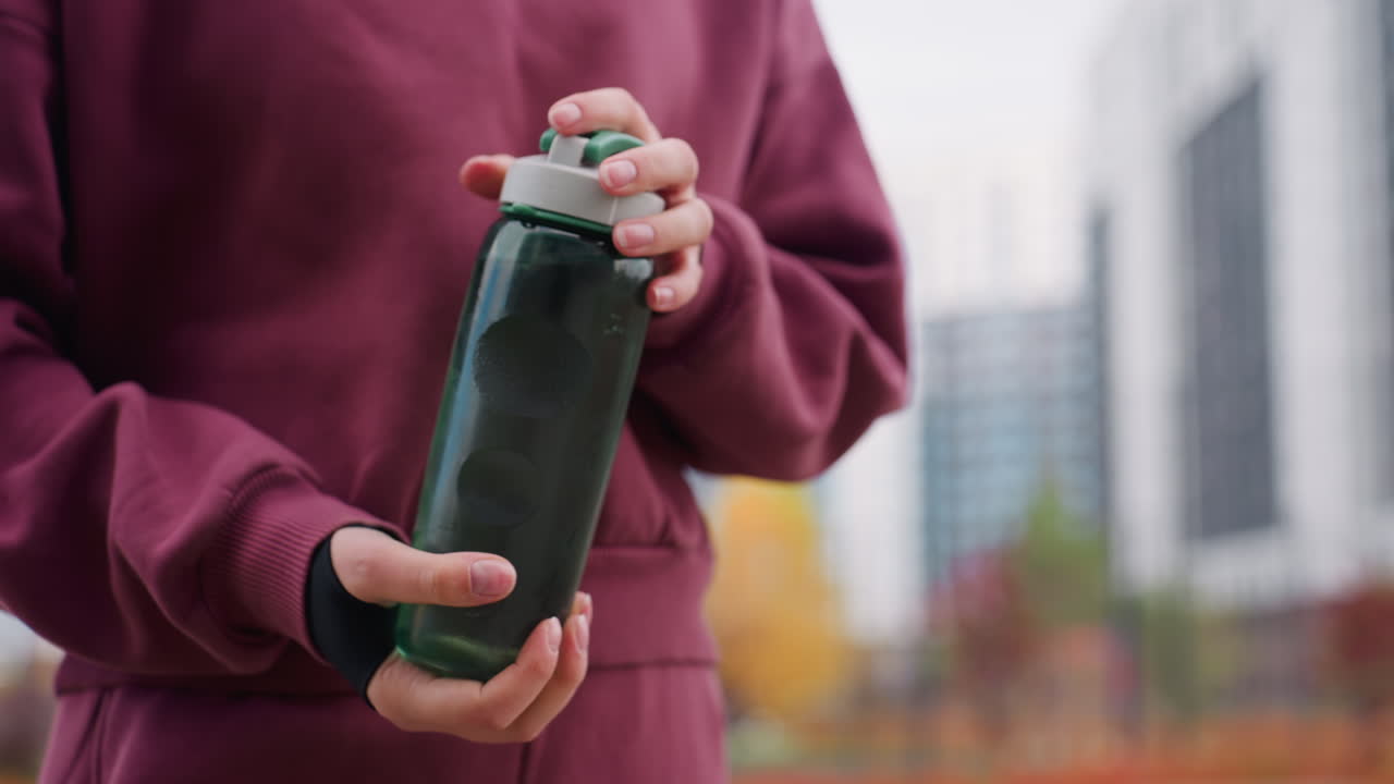Lower view of gym trainer in maroon hoodie shaking water inside bottle while walking along urban pavement with blurred high rises and spring foliage capturing fitness hydration action outdoors