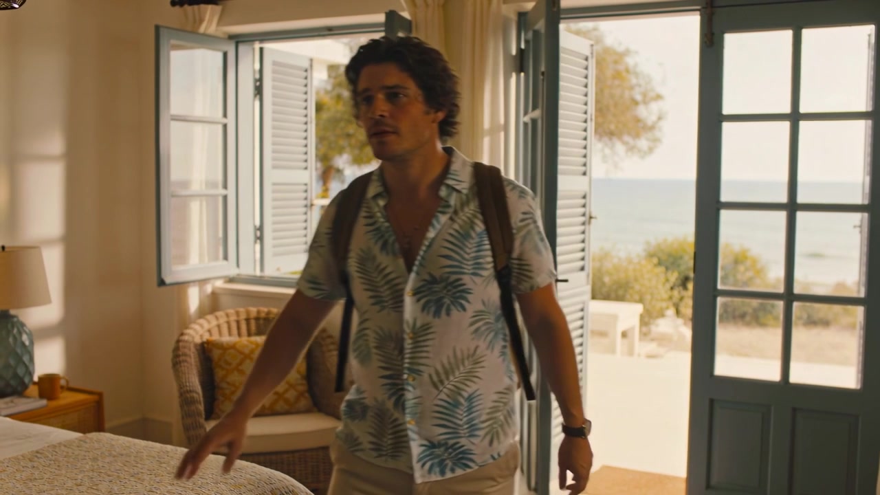 Aprendo porte blu, un uomo in camicia tropicale entra in una stanza sul mare, guardandosi intorno per appoggiare lo zaino.