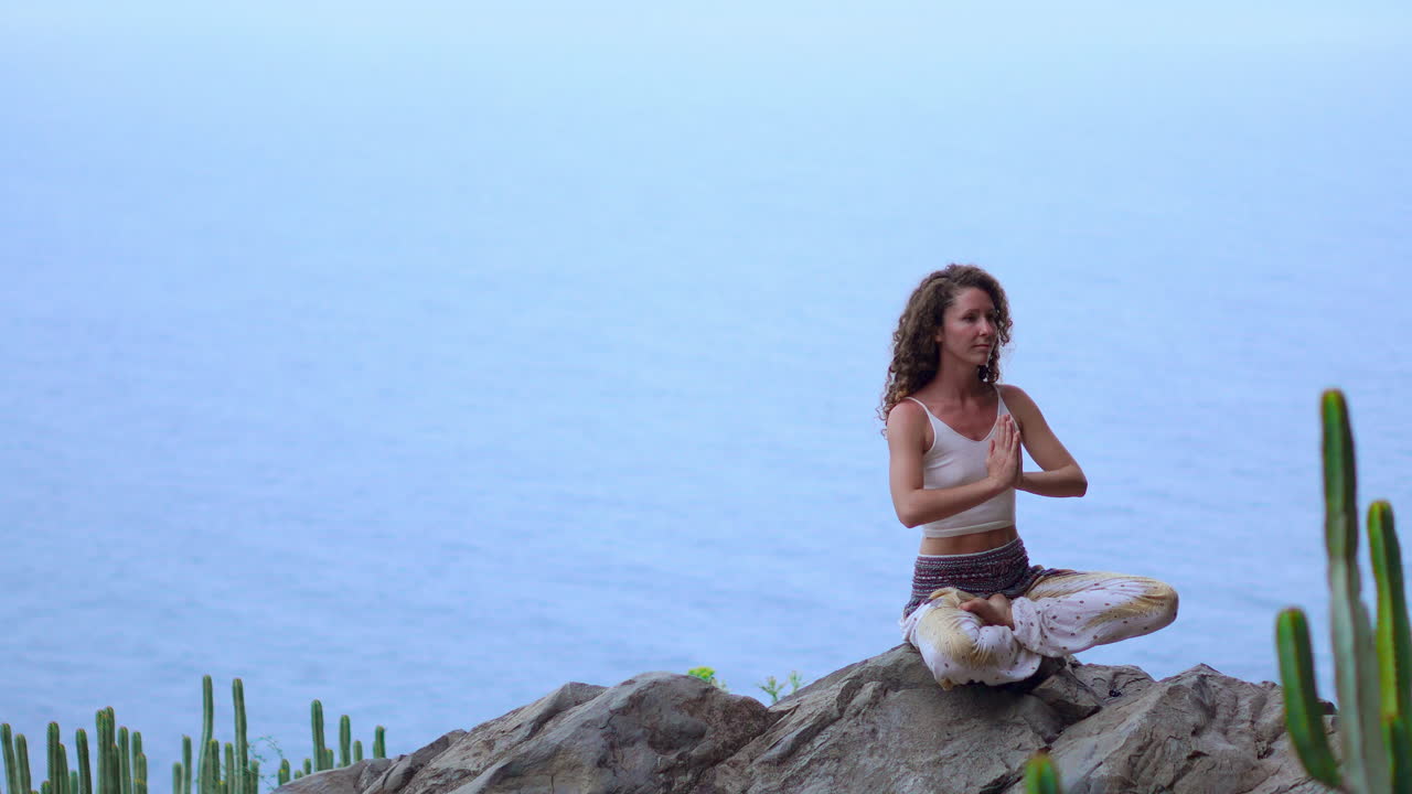 On an island's mountains, a young woman sits on a mountaintop rock, meditating in Lotus position, embracing yoga with ocean views