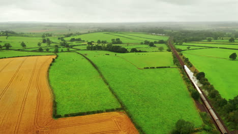 Toma-Aérea-Siguiendo-Un-Tren-De-Vapor-Con-Humo-Blanco-Que-Viaja-Por-La-Campiña-Inglesa,-Día-Nublado