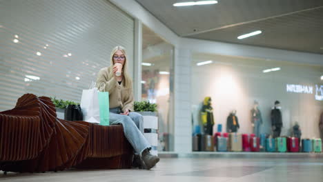 young lady sipping beverage seated on a decorative bench in a mall with shopping bag beside her, background shows store with mannequins and traveling bags displayed