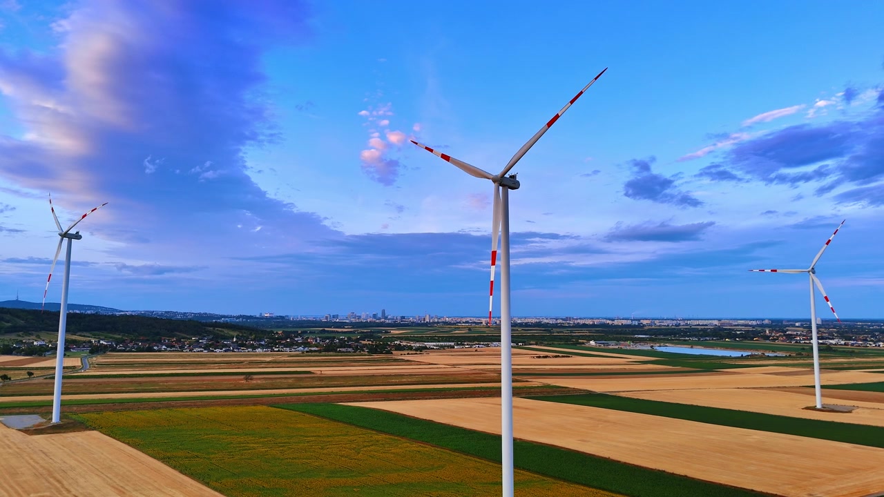 Tre turbine eoliche sopra terreni agricoli di fronte a uno skyline cittadino distante. La vista aerea mostra torri bianche con marcature rosse sulle pale, campi rettangolari, un bacino idrico e nuvole blu.