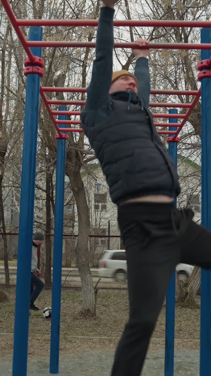 A coach jumps up and grabs a red iron bar, moving along it as he swings, in the background, a boy plays football, and cars pass by, with residential buildings and bare trees visible across the road
