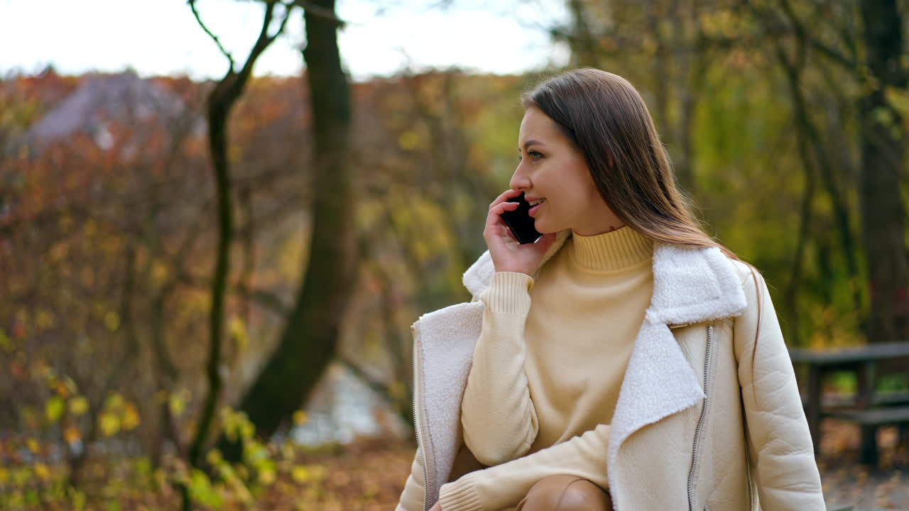Relaxed smiling lady sitting in the autumn park speaking on the phone. Brunette young woman having phone conversation outdoors. Blurred backdrop.