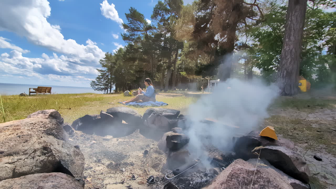 mujer joven sentada en el campamento por el fuego del campamento hoyo de humo, en la naturaleza cerca del bosque