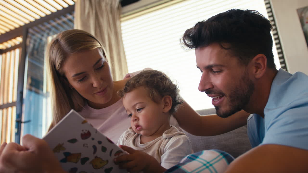 Família sentada no sofá em casa com os pais lendo um livro com o filho pequeno