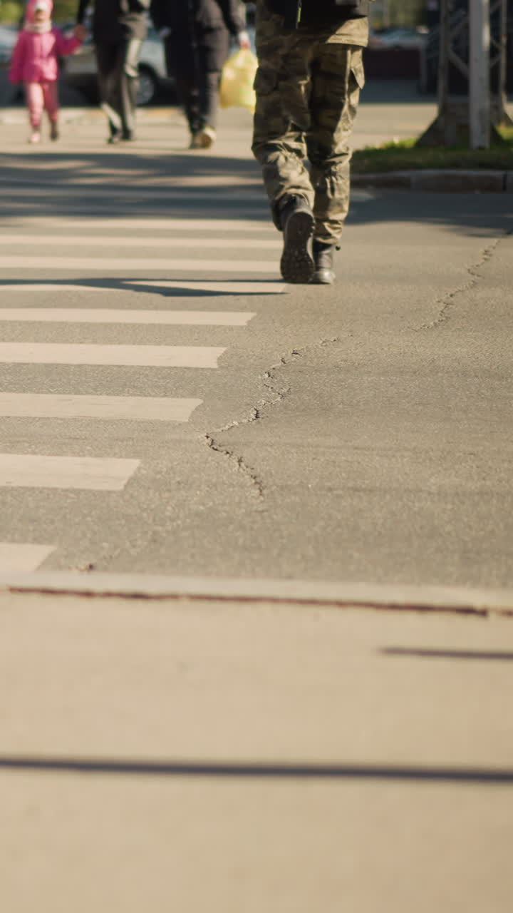 person in camouflage pants crossing street purposeful step in heavy boots, urban crosswalk with cracked asphalt markings, shoppers and children in background, steady march suggesting patrol