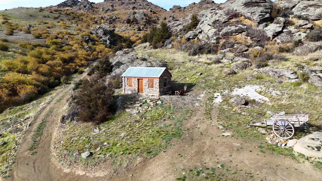 Aerial footage captures a solitary cabin amidst rocky terrain and sparse vegetation under bright daylight in Cromwell, New Zealand