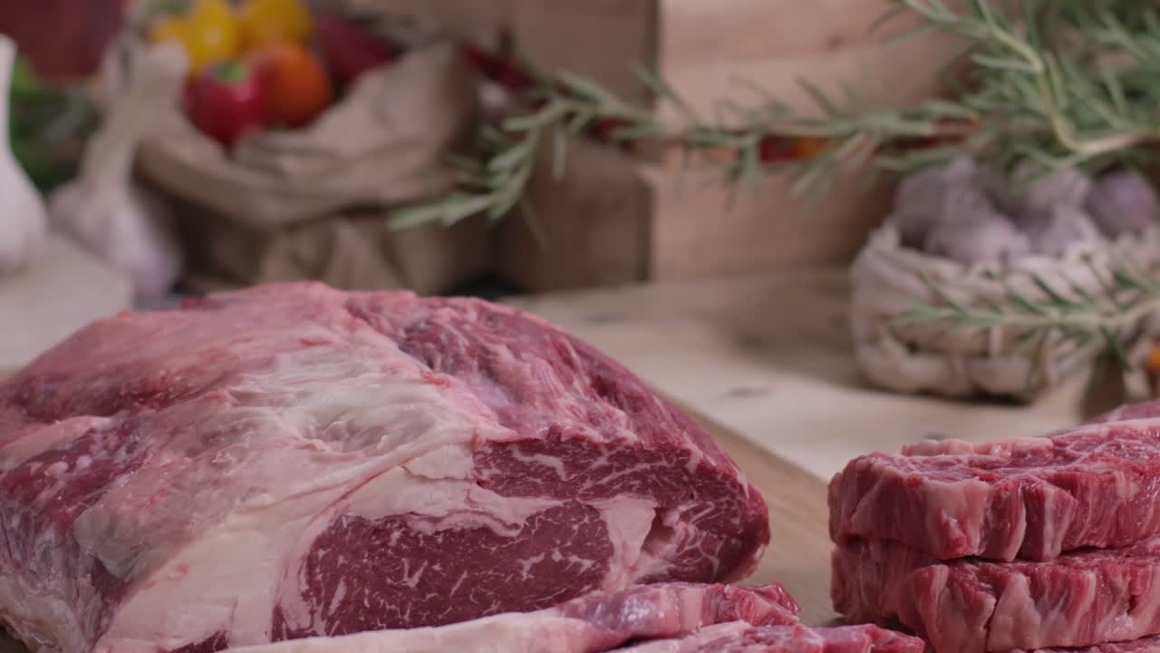 Gerakan lambat menggeser wajan di atas steak ribeye mentah dengan marbling sempurna dan daging panggang sapi besar di atas papan kayu, ditata untuk pemotretan makanan komersial dengan herba dan sayuran segar