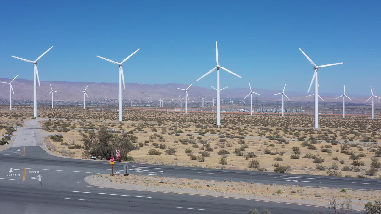 Many white renewable windmill turbines in arid countryside field with rotating propellers from offshore wind generating energy on bright blue sunny sky day, pan left