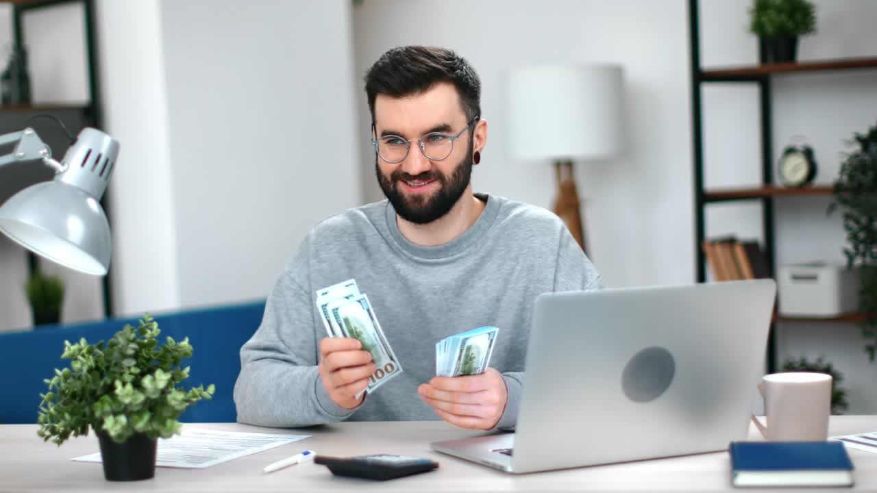 Freelancer emocionado revisando dinero en efectivo (dólares) ganando dinero por internet en su portátil de trabajo