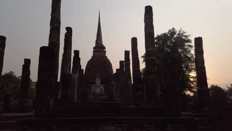 Push-in-towards-large-Buddha-seated-statue-between-historic-stone-column-ruins-in-religious-garden-site-attraction-at-sukhothai,-thailand