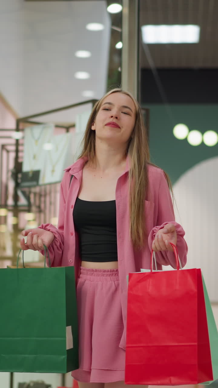 hermosa dama de vestido rosa toma una respiración profunda, levanta sus bolsas de compras rojas, verdes y de menta mientras está de pie frente a una tienda, luego transfiere la bolsa verde a su mano derecha mientras se aleja