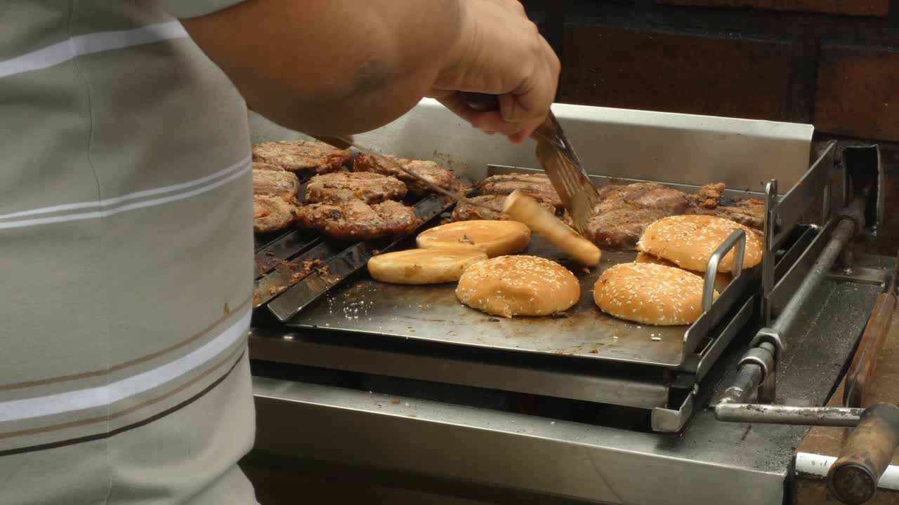 A person flipping burger buns as they toast on the flattop part of the barbecue grill. On the left the burgers are cooking.