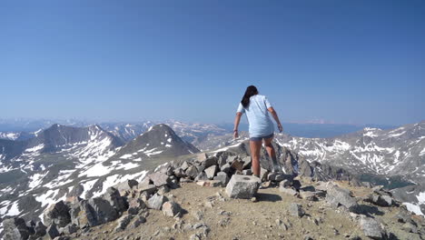 back view of young woman atop of peak with stunning view of mountain range on sunny day