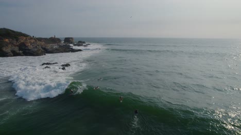 Rough-sea-with-waves,-surfers-surfing-next-to-rocks