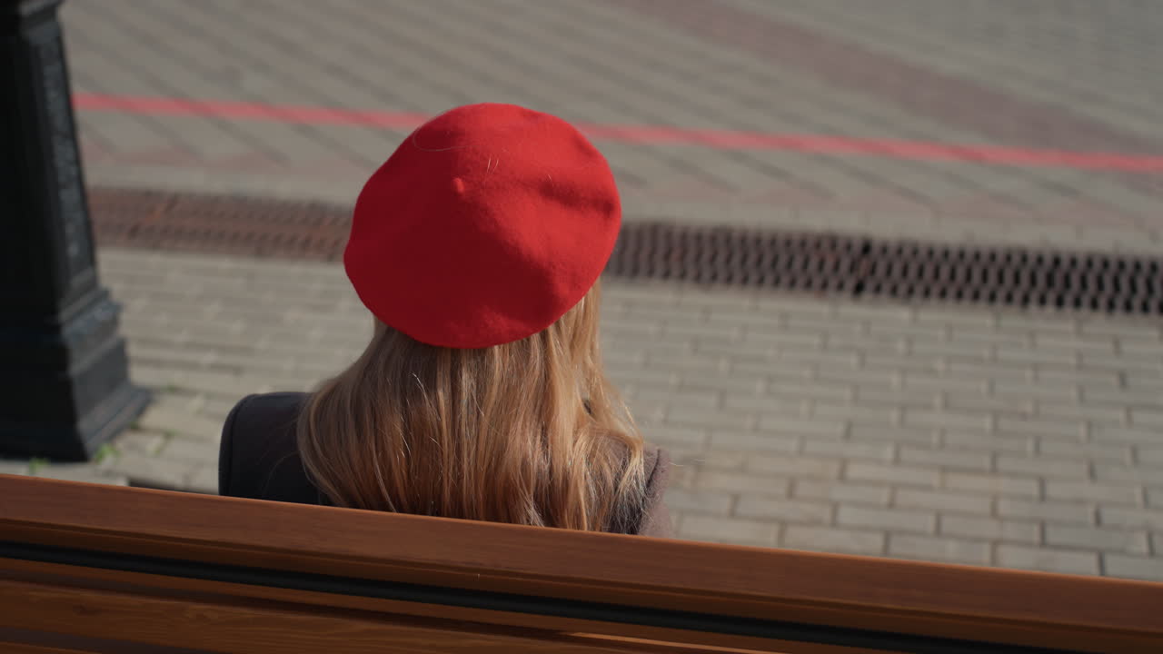 Kvinde hviler på bænk på rolig byplads. Opmærksom kvinde med smartphone sidder i et fredfyldt bymiljø. Kvinde med rød baret og brun frakke slapper af på bænk på en fredelig byplads