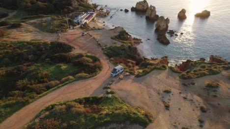 Luftaufnahme-Eines-Umkreisenden-Wohnmobils,-Das-Auf-Der-Felsigen-Küstenklippe-Von-Praia-Dos-Arrifes-Mit-Blick-Auf-Die-Malerische-Meereslandschaft-Bei-Sonnenaufgang-Am-Atlantischen-Ozean-Geparkt-Ist