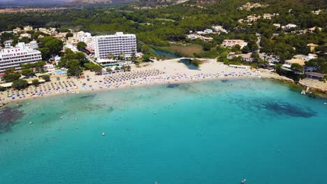 Toma-Aérea-De-La-Playa-De-Canyamel-Con-Aguas-Turquesas-Y-Una-Playa-Llena-De-Gente-Durante-El-Verano,-En-Mallorca,-España,-En-El-Mar-Mediterráneo.