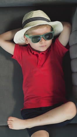 little boy in straw hat and sunglasses relaxes on couch moving with bare foot. stylish toddler rests in hotel room after arrival upper close view